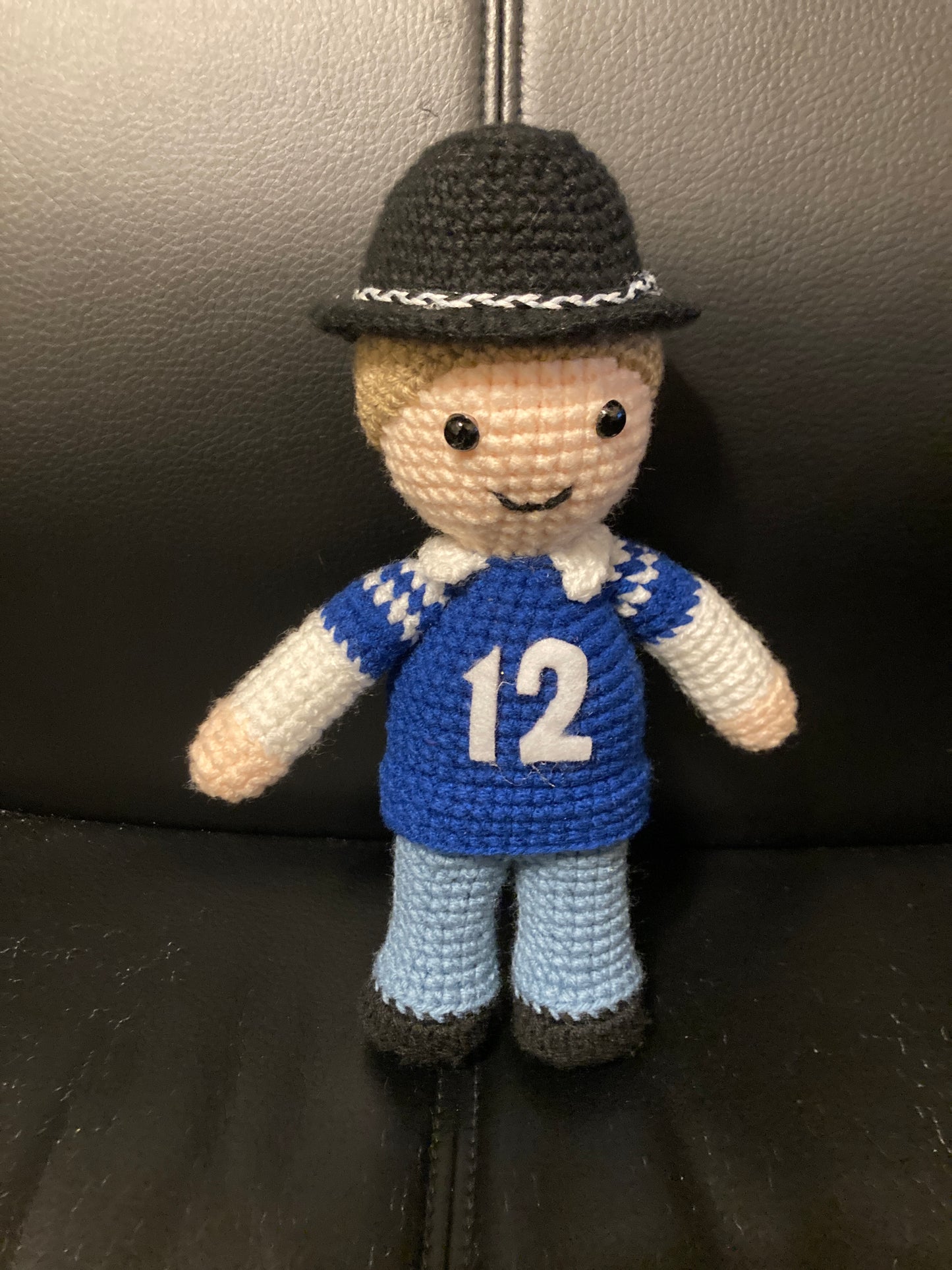 A crochet doll with a blue jersey number 12, blue pants, and a black hat.